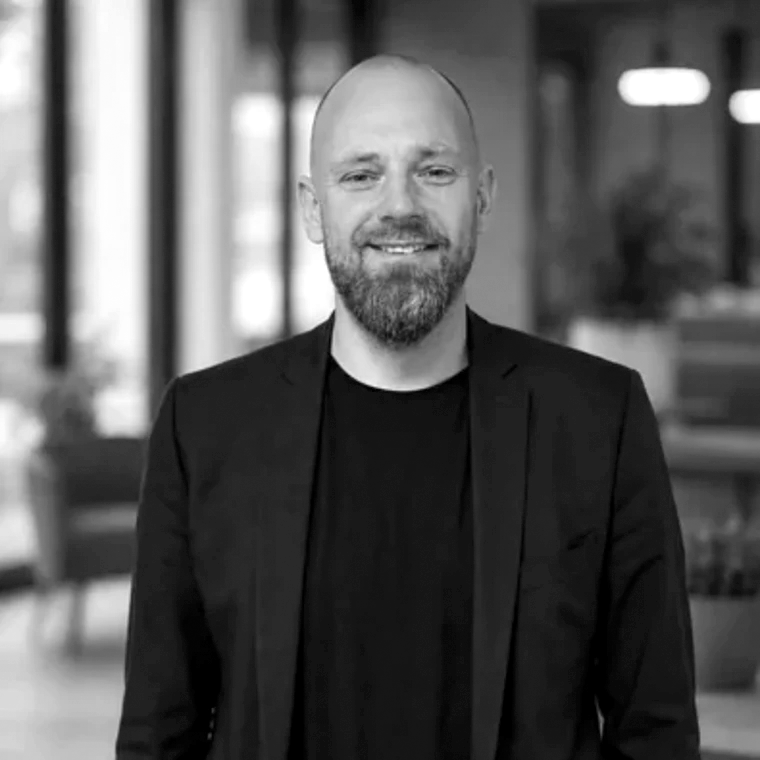 Sort/hvidt portræt af smilende mand med kort skæg og barberet hoved i mørk blazer og T-shirt foran et sløret kontorlandskab