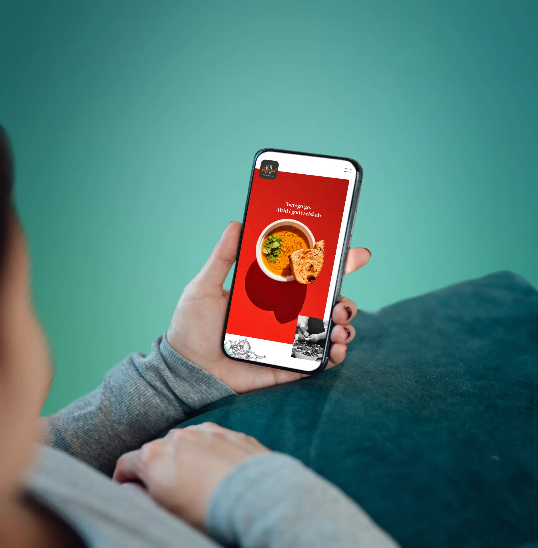 Person holder en smartphone med Værsgo' Go på skærmen foran en teal væg og et mørkegrønt pude