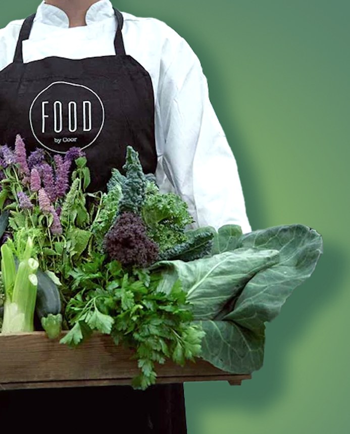 Kok i hvid jakke og sort forklæde med FOOD by Coor-logo bærer en trækasse med kål, persille og blandede urter