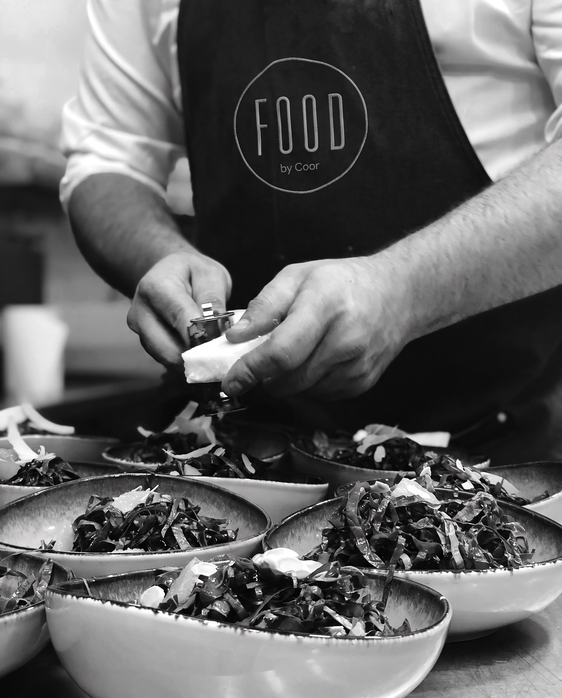 Kok i hvide ærmer og sort forklæde med FOOD by Coor-logo skaver hvid topping over en række skåle med retter