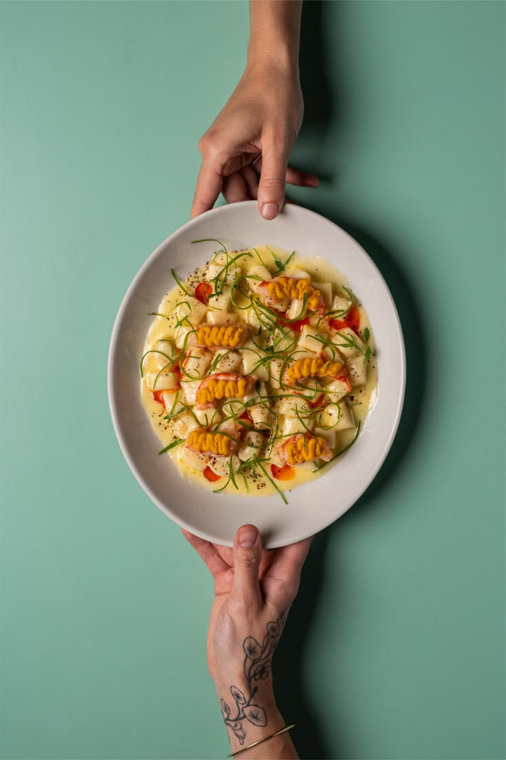 Hænder holder en skål med gnocchi i krebet sauce, rejer og uni mod en mintgrøn baggrund