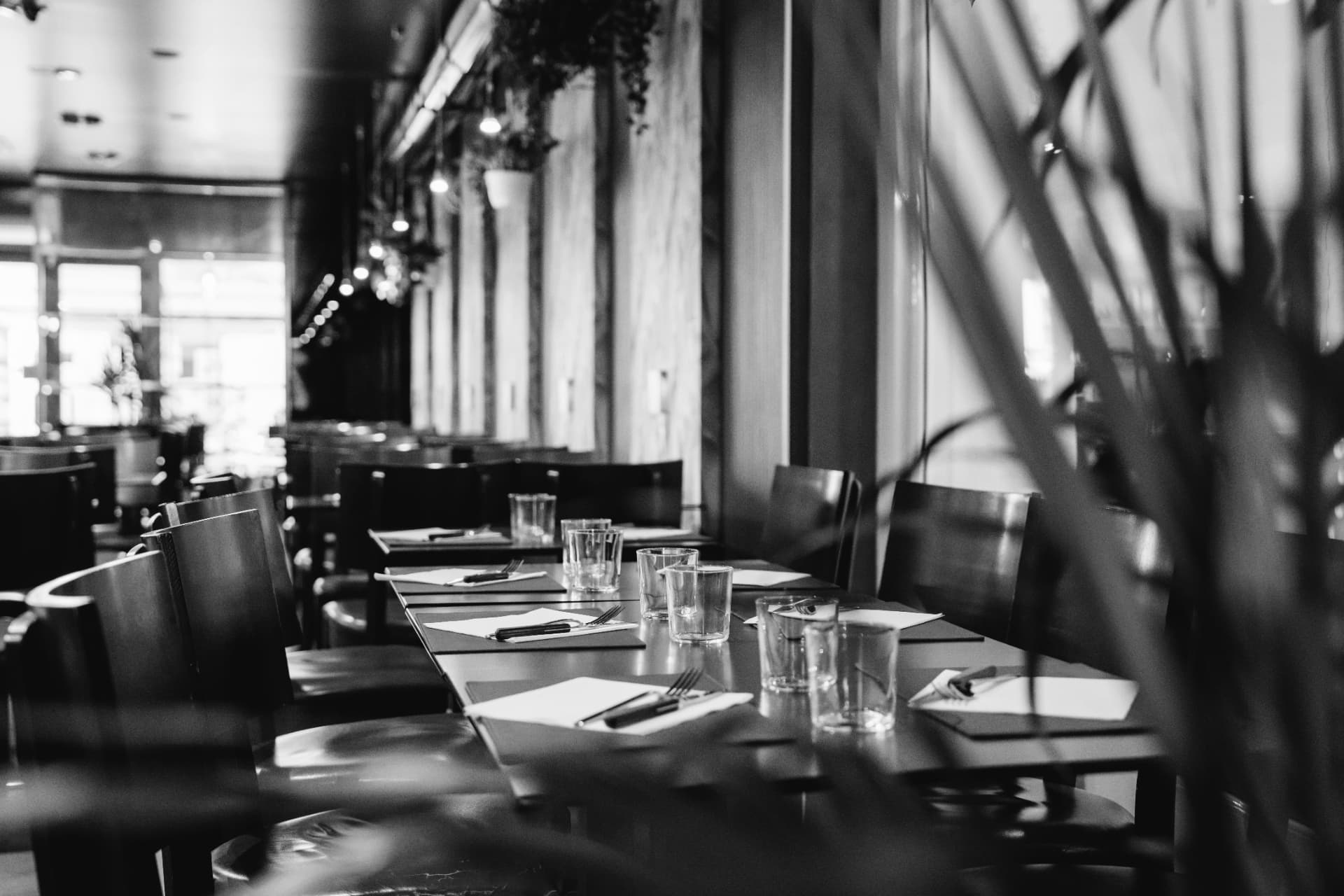 Sort/hvidt foto af et restaurantlokale med borde dækket med glas og bestik i sollys fra vinduerne