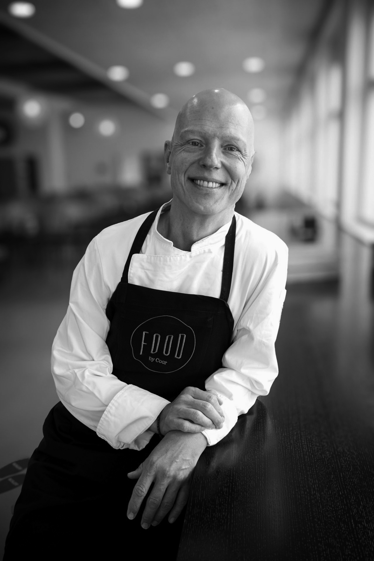 Sort/hvidt portræt af en smilende kok i hvid kokkejakke og sort FOOD by Coor-forklæde, der læner sig mod en disk