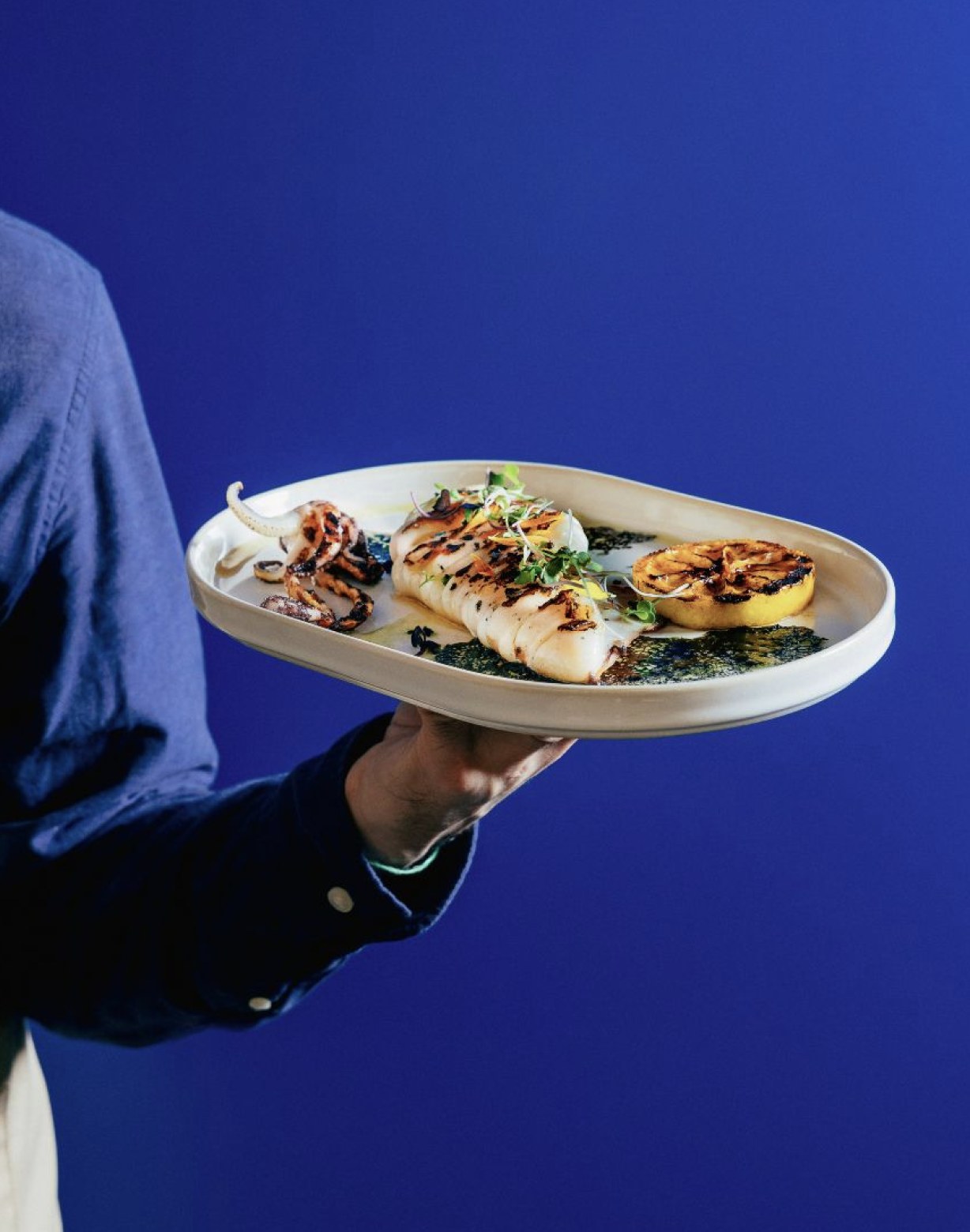 En hånd præsenterer et fad med grillet fisk og blæksprutte samt grillet citron mod en dyb blå baggrund